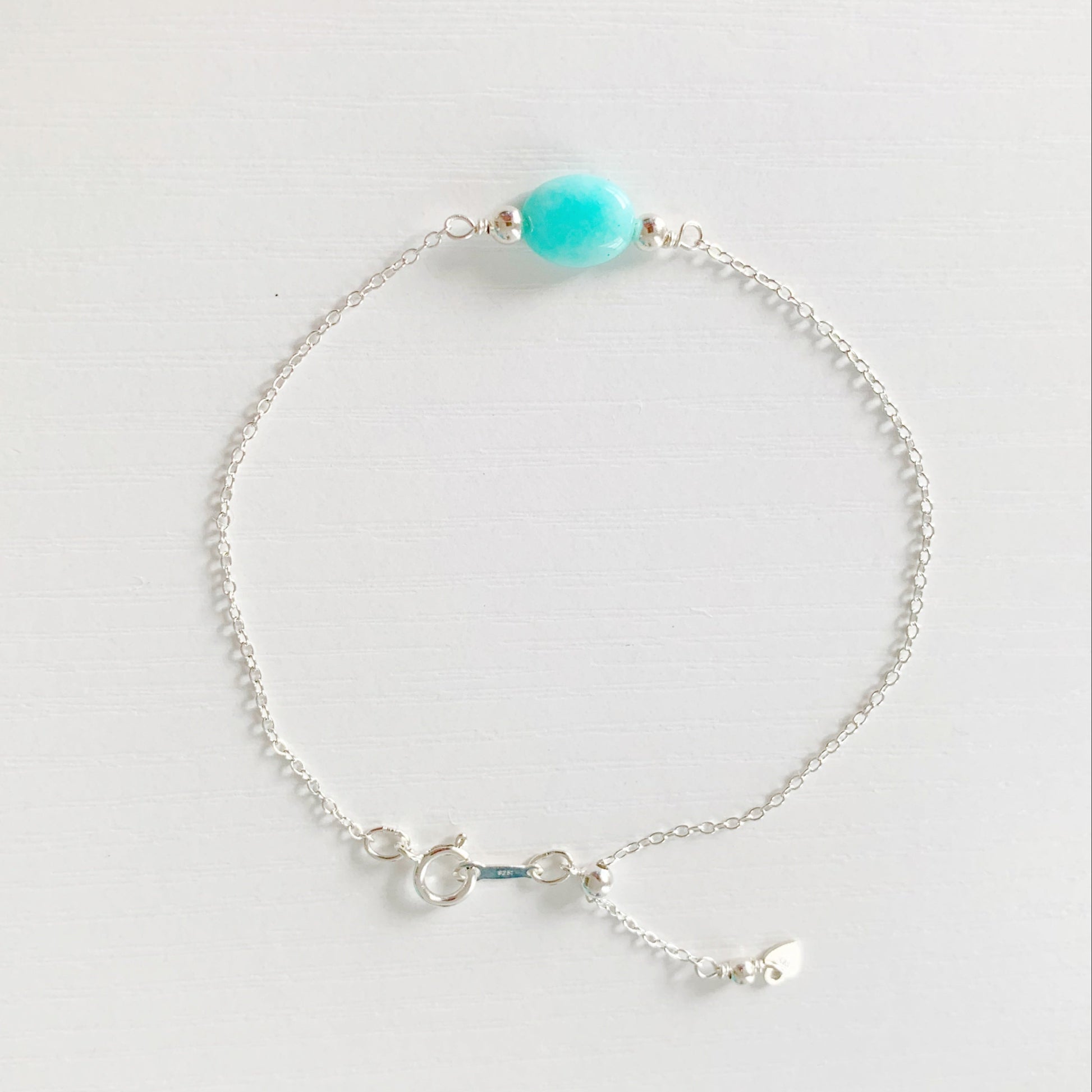the laguna bracelet by mermaids and madeleines is a sterling silver chain based bracelet with an aqua amazonite oval bead a the center and with sterling slide bead near the clasp making it adjustable. this bracelet is photographed from the top down on a white surface