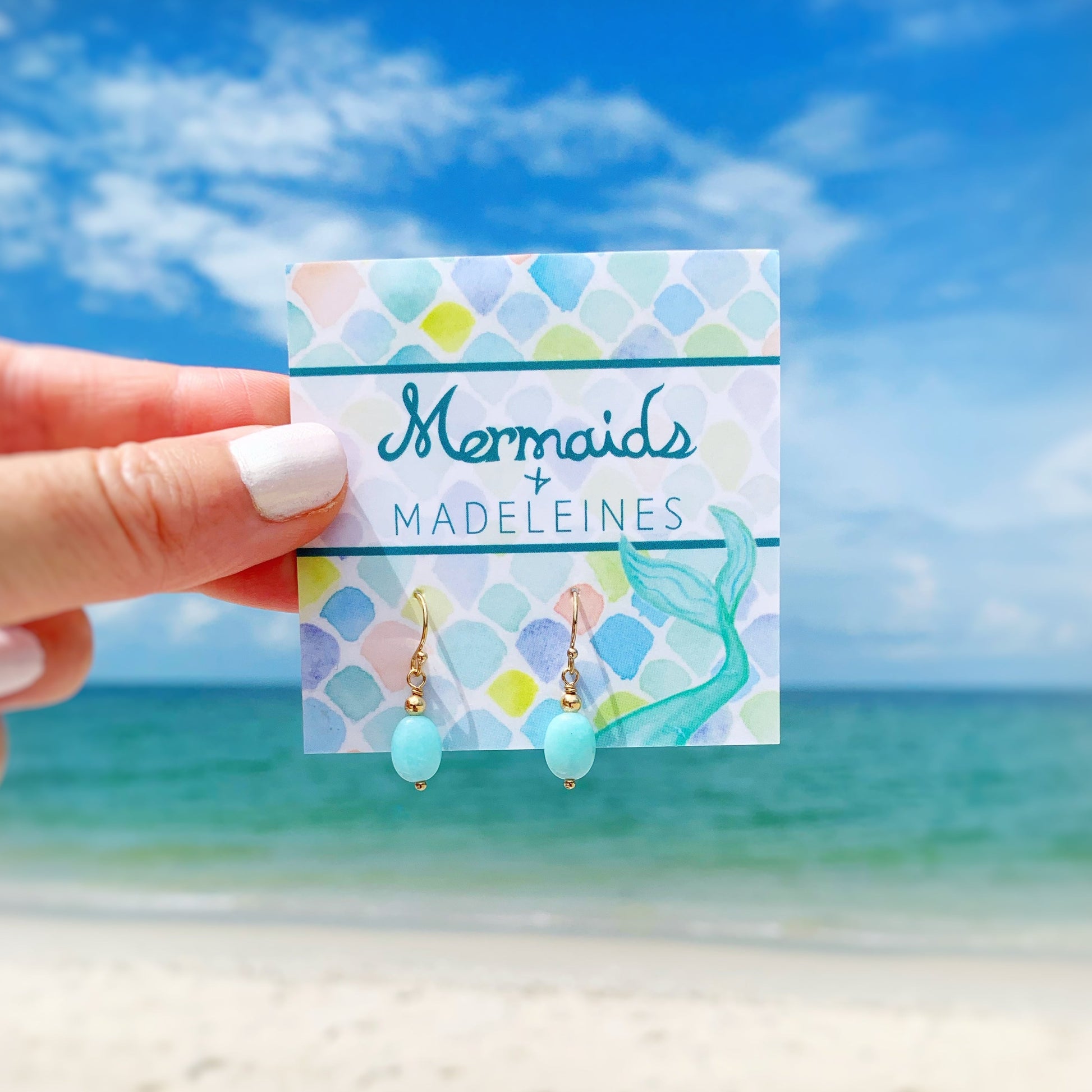 Laguna Earrings by mermaids and madeleines are aqua amazonite oval stones with 14k gold filled findings. this pair is hanging on a branded earring card and held up in front of a beach background