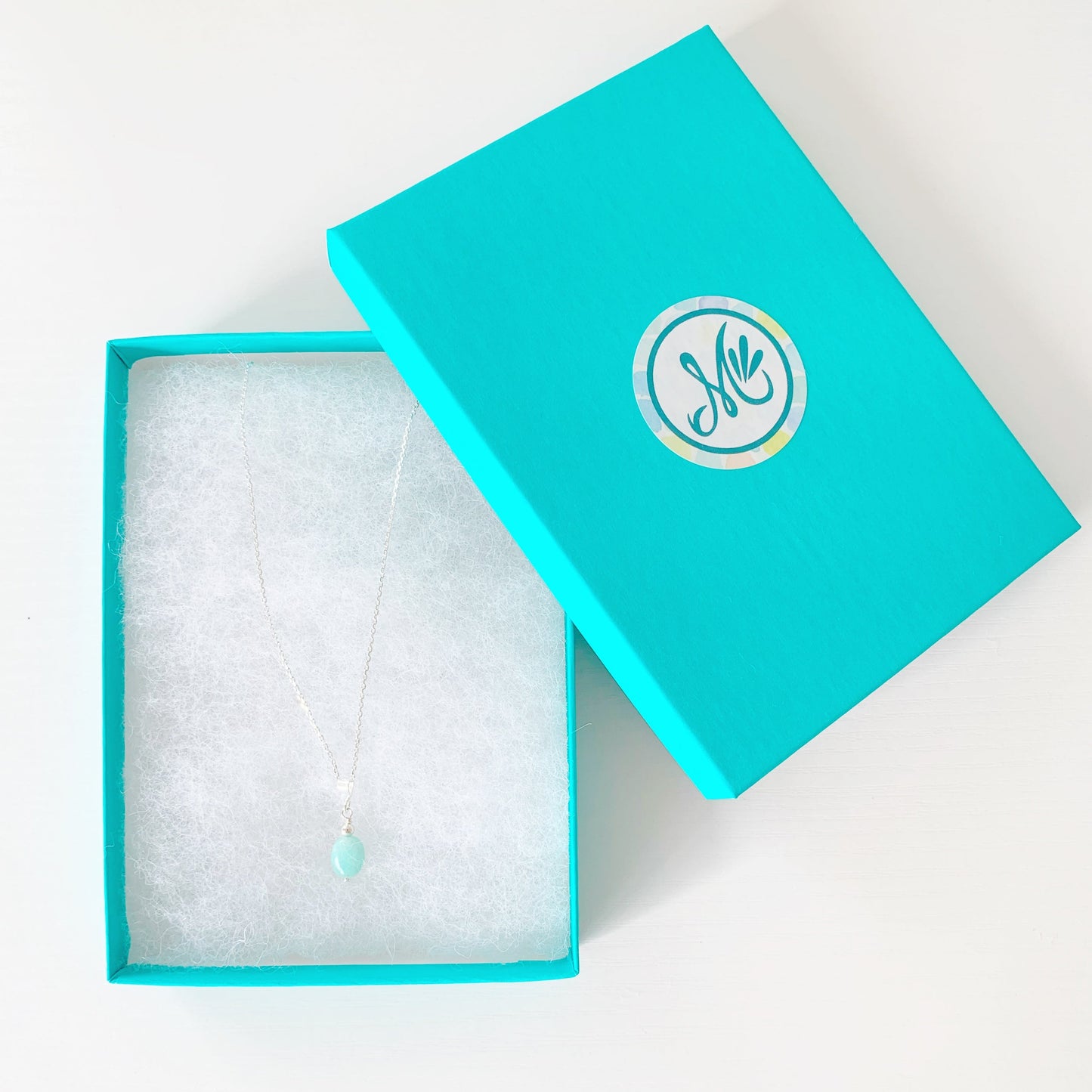 the laguna necklace by mermaids and madeleines is created in a pendant style. the necklace features a smooth, oval amazonite bead, dropped from sterling silver findings and chain. this is a photo of the laguna necklace in a teal gift box on a white surface