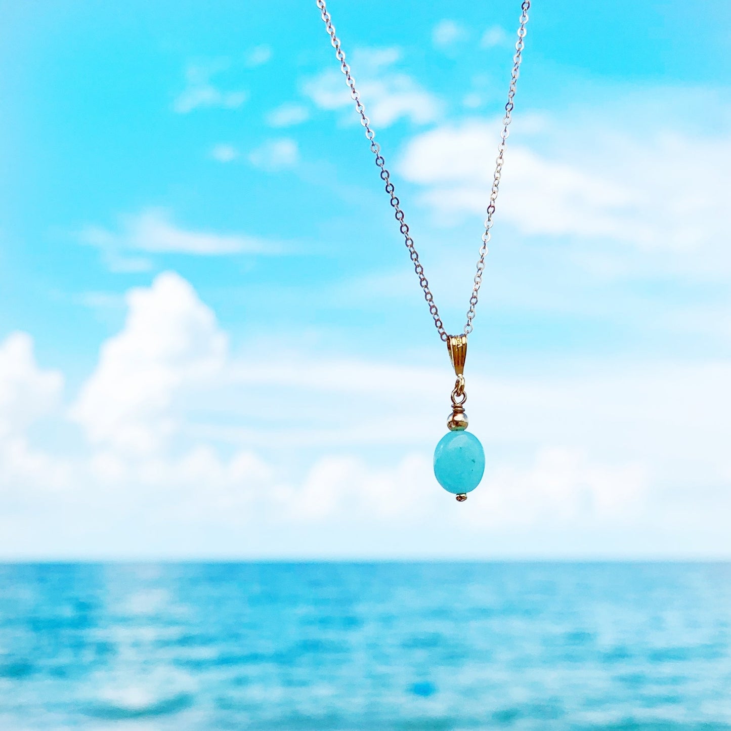 Laguna necklace by mermaids and madeleines is created with an aqua color amazonite oval stone and  hangs from a 14k gold filled chain. this necklace is pictured suspenced over a blue sky and beachy background