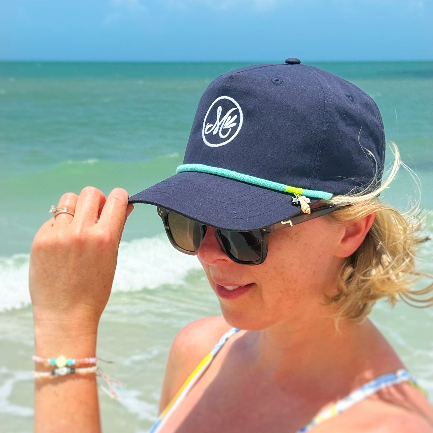 A person wearing a navy blue snapback cap with a teal cord in the front and an "M" logo with the beach in the background