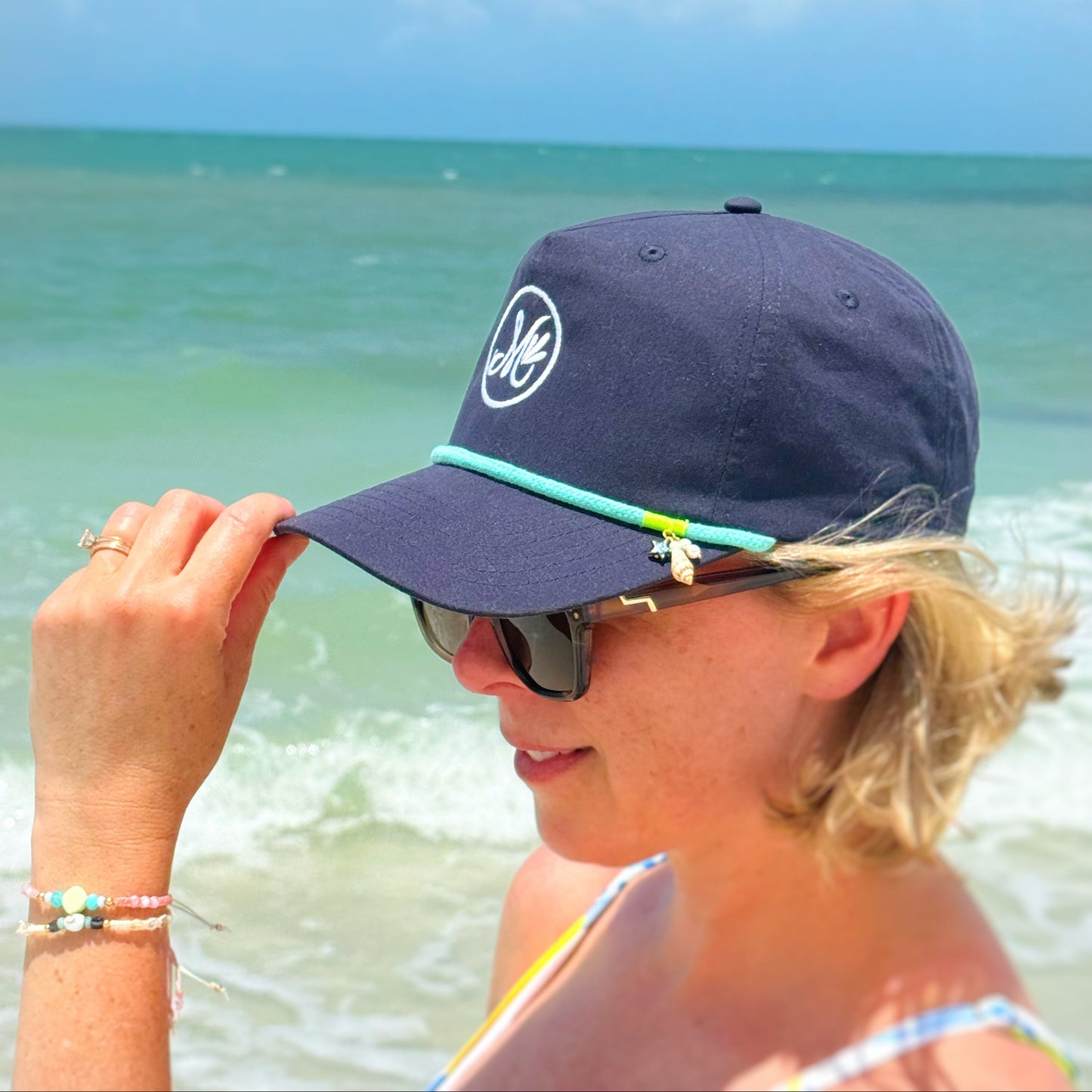 A person wearing a navy blue snapback cap with a teal cord in the front and an "M" logo with the beach in the background