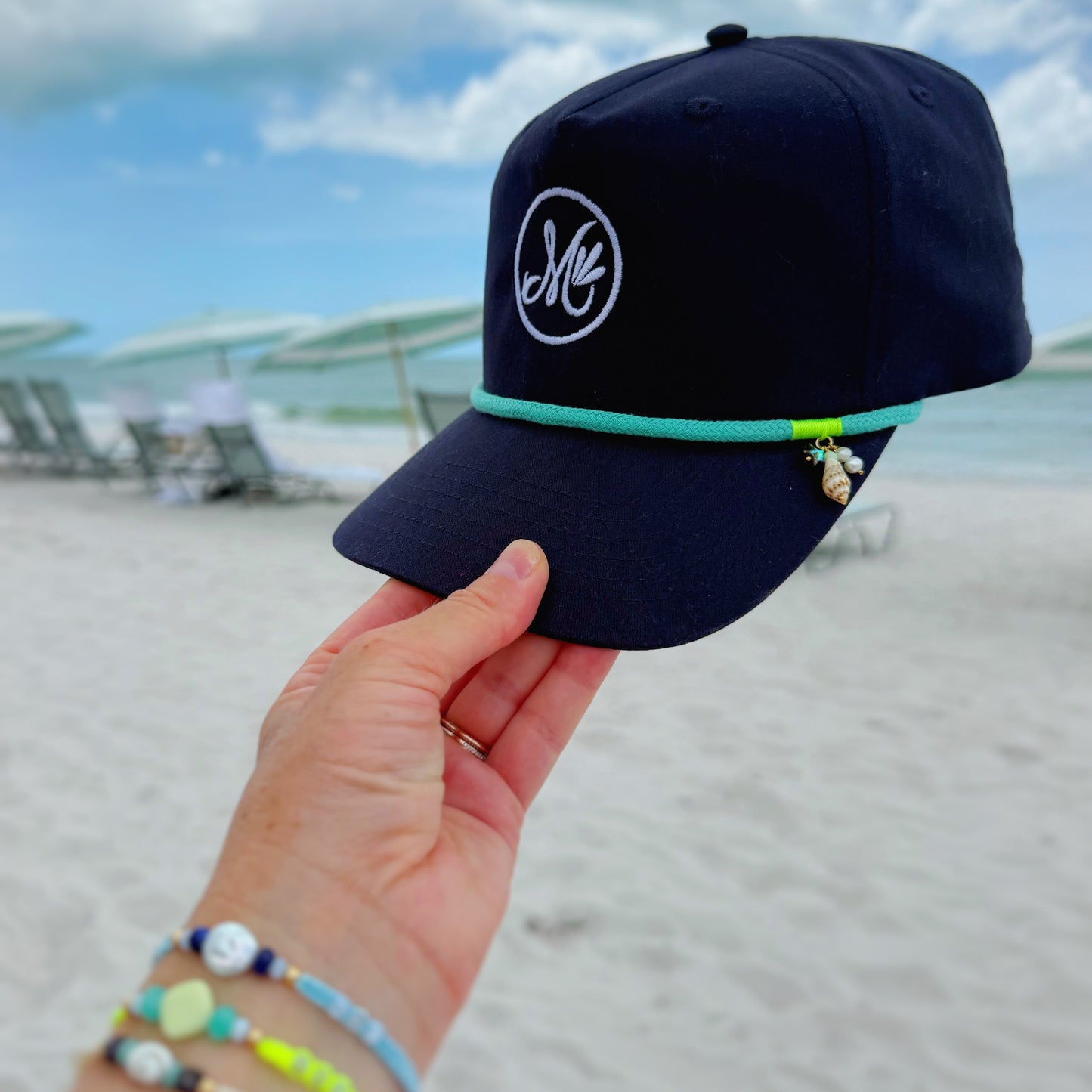 a navy blue snapback cap with a white m logo and teal rope above the bill. this hat is held in a hand with colorful bracelets on and a beach background