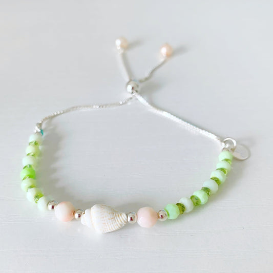 the margarita shellebration bracelet by mermaids and madeleines is a sterling silver and beaded adjustable bracelet with a shell and coral beads at the center complimented by alternating green opal and peridot. this bracelet is photographed on a white surface