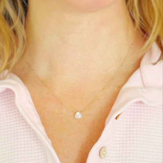 Gold necklace with a small moonstone pendant worn by a person in a light pink shirt.