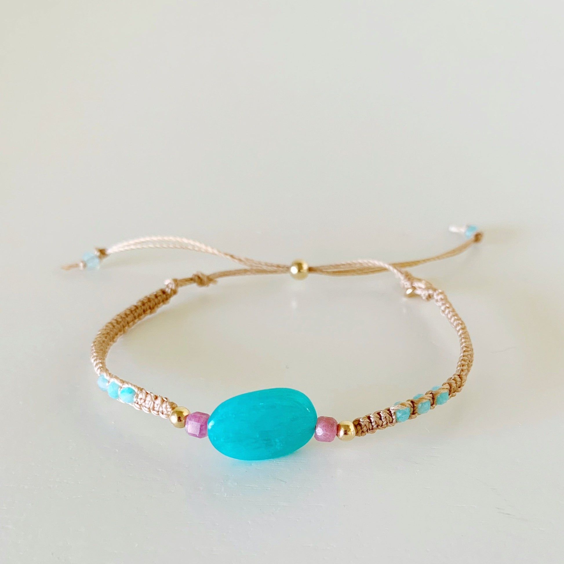 the harwich port adjustable macrame bracelet by mermaids and madeleines in color moonlit beach is created in a friendship style bracelet with light tan color cord and a slide bead clasp. this bracelet features a bright amazonite bead at the center with smaller amazonite rondelles on the sides and complimented by pink tourmaline and 14k gold filled beads. this bracelet is facing forward and is photographed on a flat white surface