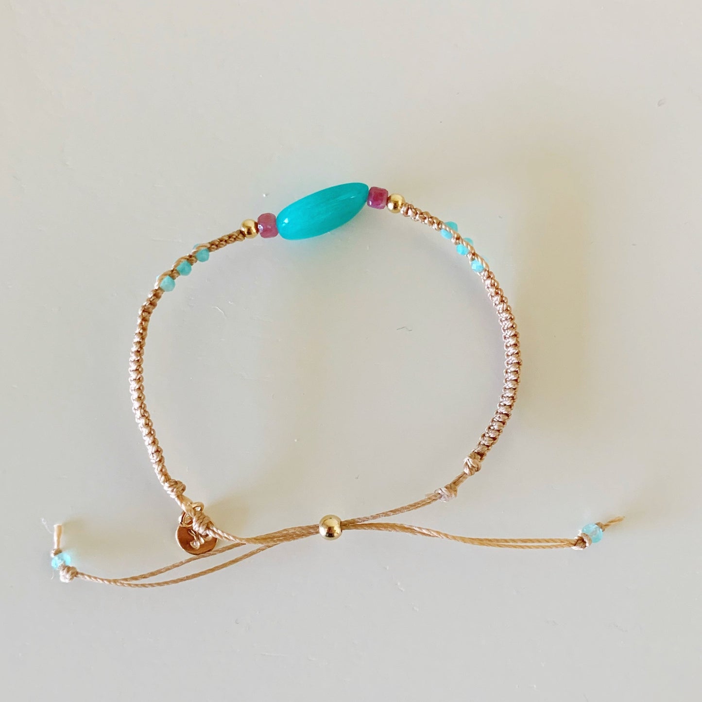 the harwich port adjustable macrame bracelet by mermaids and madeleines in color moonlit beach is a friendship style bracelet made with tan cord and a slide bead clasp. this bracelet has a large amazonite bead at center with pink tourmaline and 14k gold filled beads on either side. this image is a full view and profile picture of the bracelet photographed on a white surface