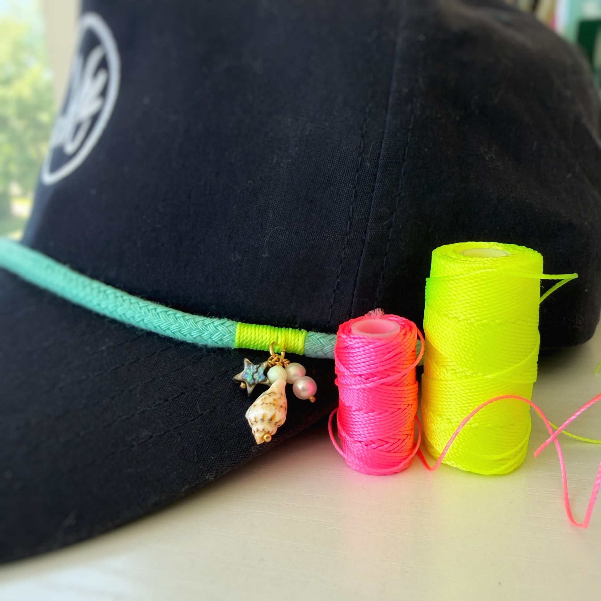 a close up side view of a navy blue snapback cap with a teal rope above the bill and an m logo in white. in front of the hat are spools of neon cord on a white surface