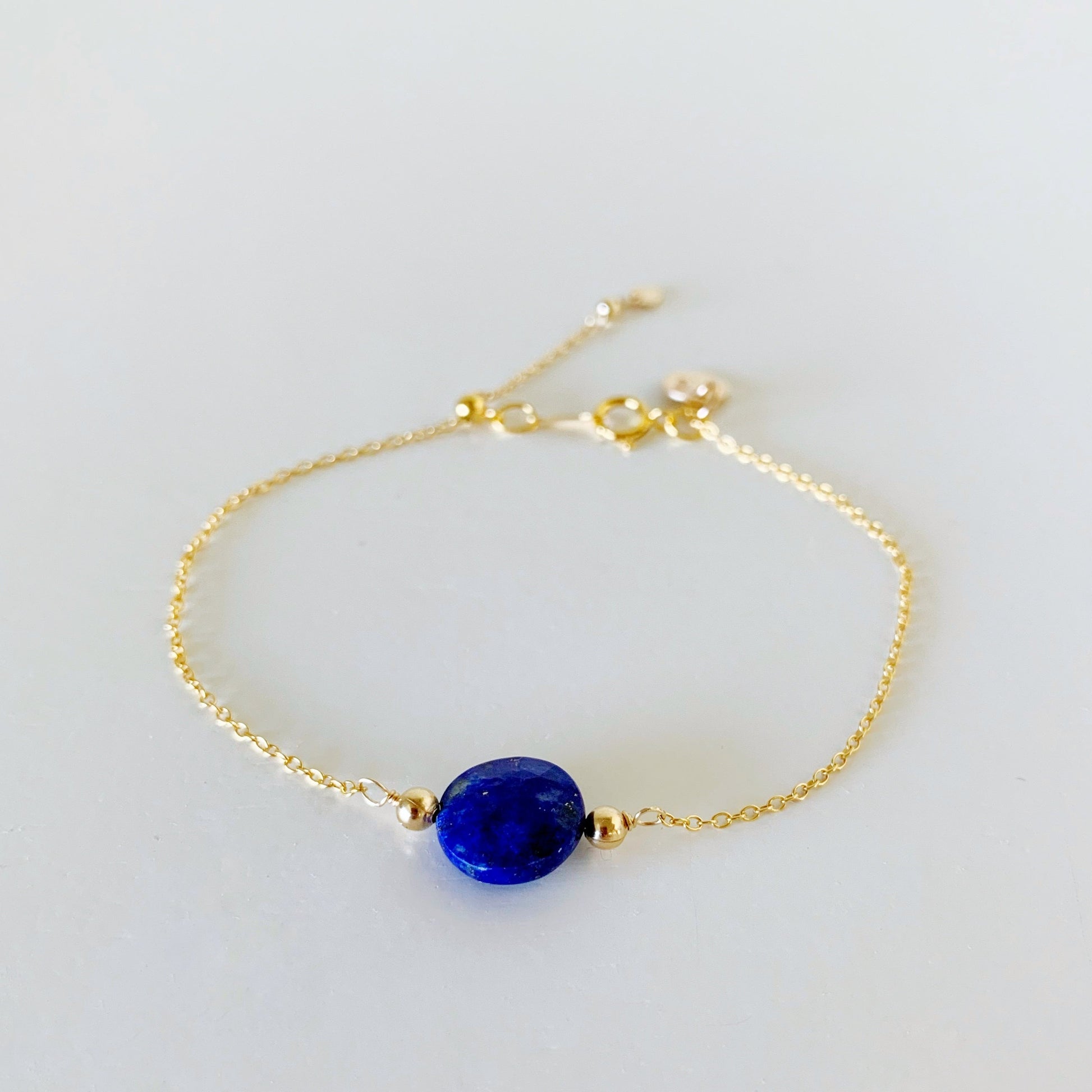 the neptune adjustable bracelet by mermaids and madeleines is a 14k gold filled chain based bracelet with a silicone slide bead at the clasp. theres also a royal blue lapis faceted coin bead at the center of the bracelet. this piece is photographed on a white surface