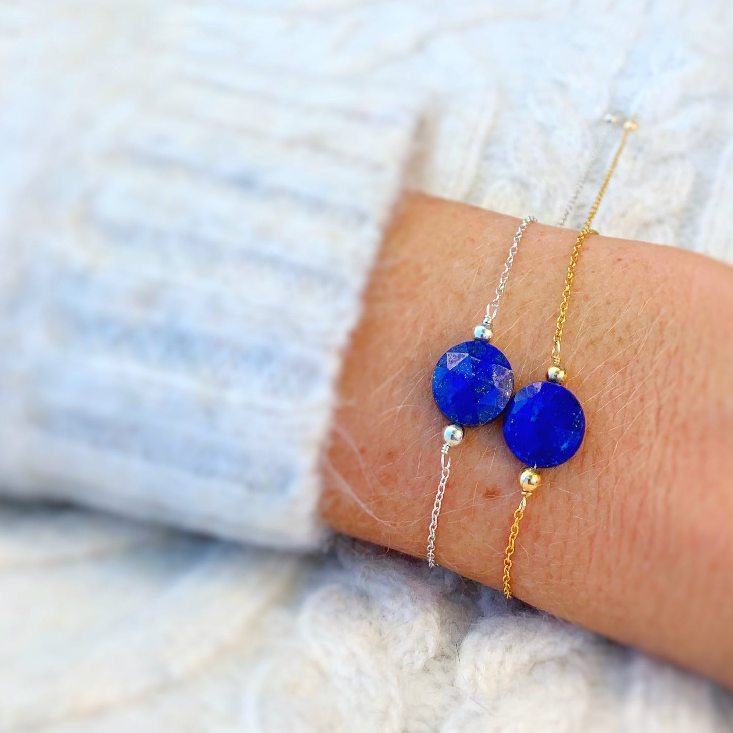 the mermaids and madeleines neptune bracelet is an adjustable chain bracelet available in sterling silver or 14k gold filled. the bracelet has a lapis coin bead at the center with a slide bead at the clasp. this is a picture of a person's wrist with a sterling silver neptune bracelet and a 14k gold filled neptune bracelet worn on it with a sweater on the upper arms and the bracelets photographed on the wrist