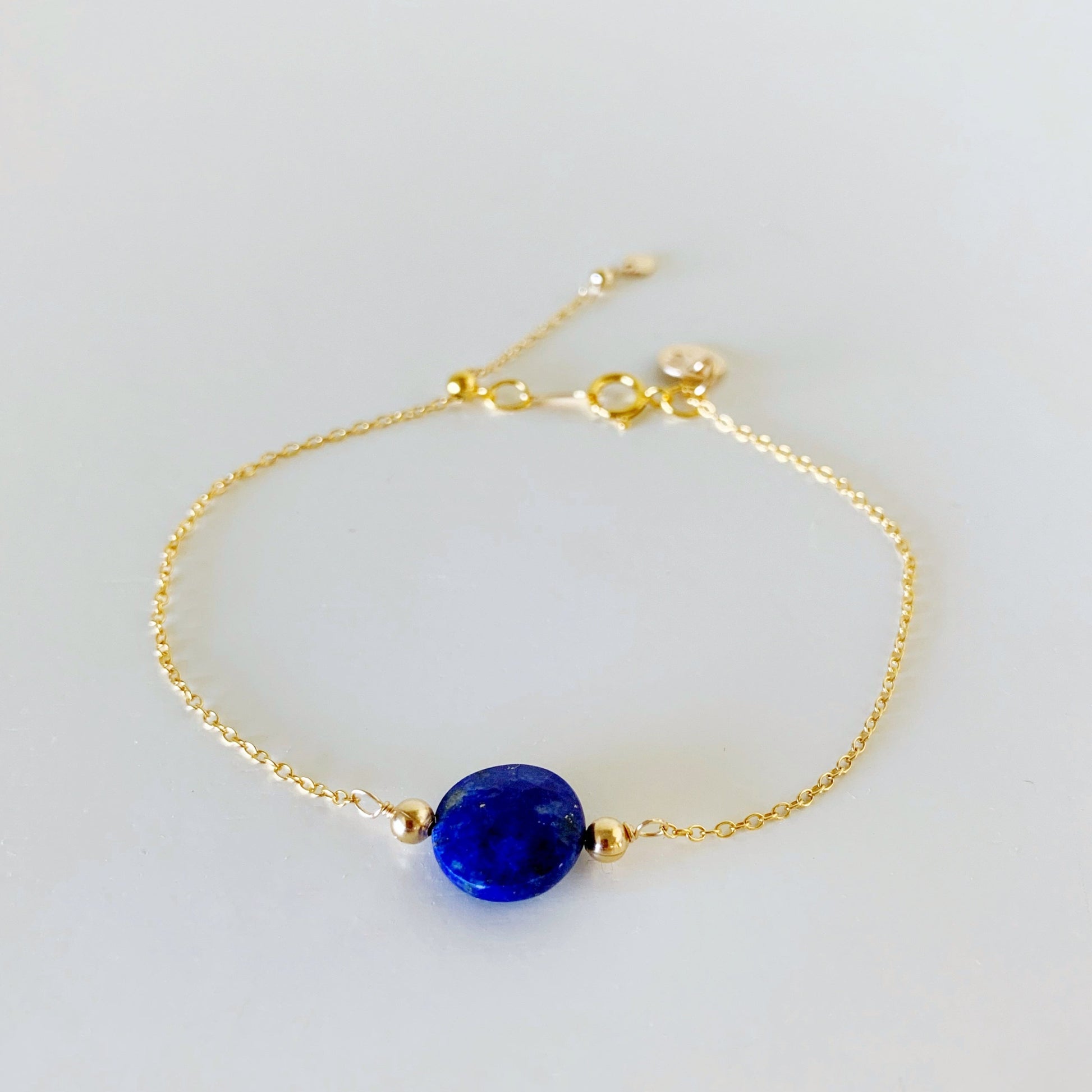 the neptune adjustable bracelet by mermaid and madeleines is a 14k gold filled chain based design with a slide bead at the clasp. there's also a royal blue lapis faceted coin bead at the center. this piece is photographed from the front on a white surface