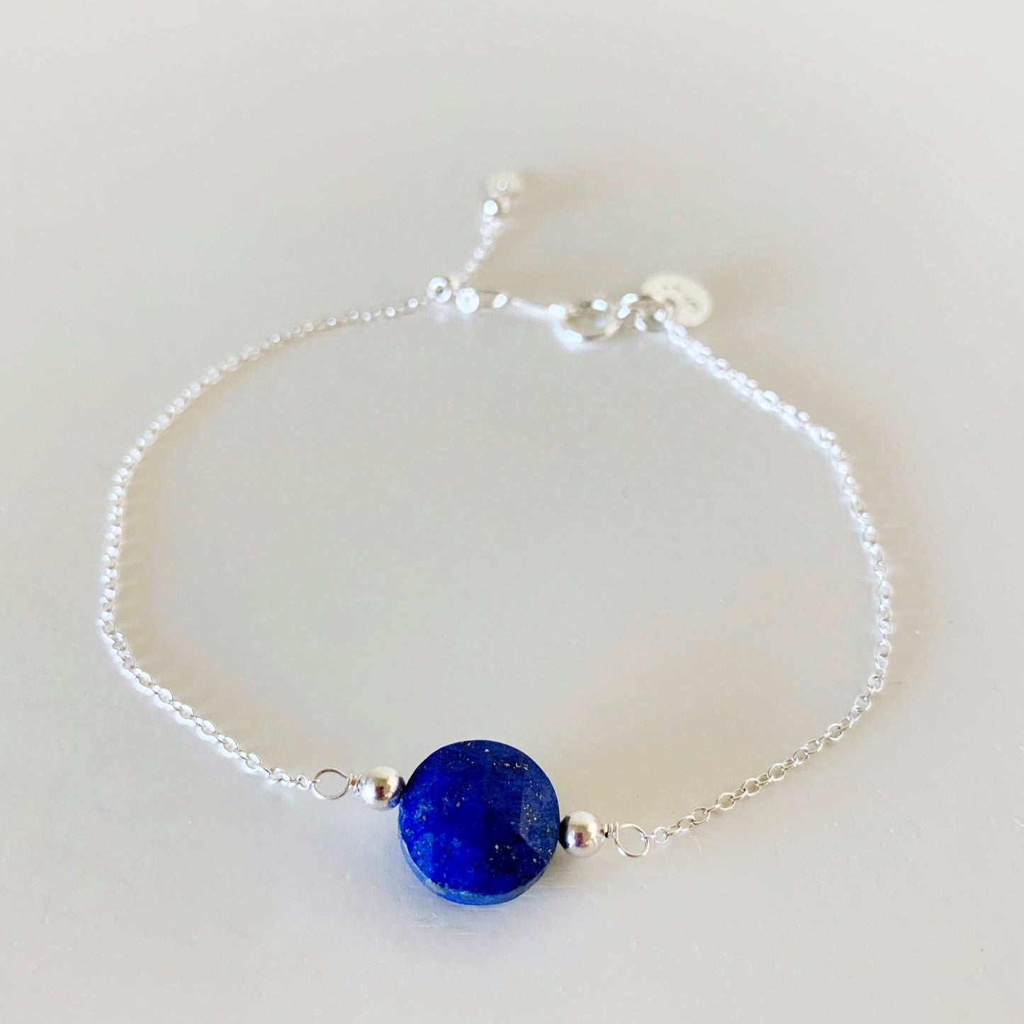 the neptune bracelet by mermaids and madeleines is a sterling silver chain based bracelet with an adjustable slider bead at the clasp. theres a royal blue faceted coin lapis bead at the center of the bracelet. this one is photographed from the front and flat on a white surface