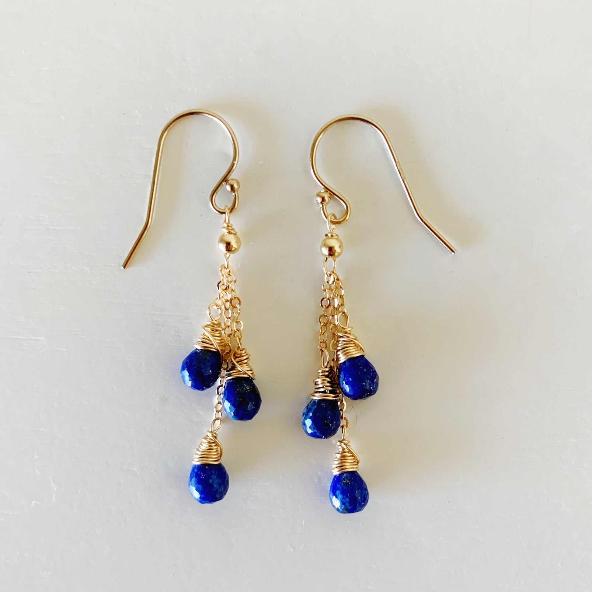 the mermaids and madeleines neptune earrings are created with 3 strands of 14k gold filled chain on each hook earring with a lapis teardrop bead wire wrapped at the bottom of each strand. this pair of earrings is photographed flat on a white surface