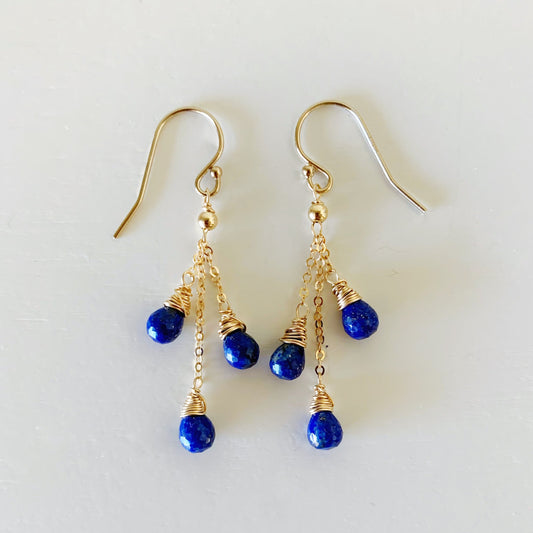 the neptune earrings by mermaids and madeleines are a dangle style earrings with 3 tear drop lapis beads dropped on each earring suspended by 14k gold filled chain from an earring hook. this pair of earrings is photographed flat with the strands splayed out on a white surface