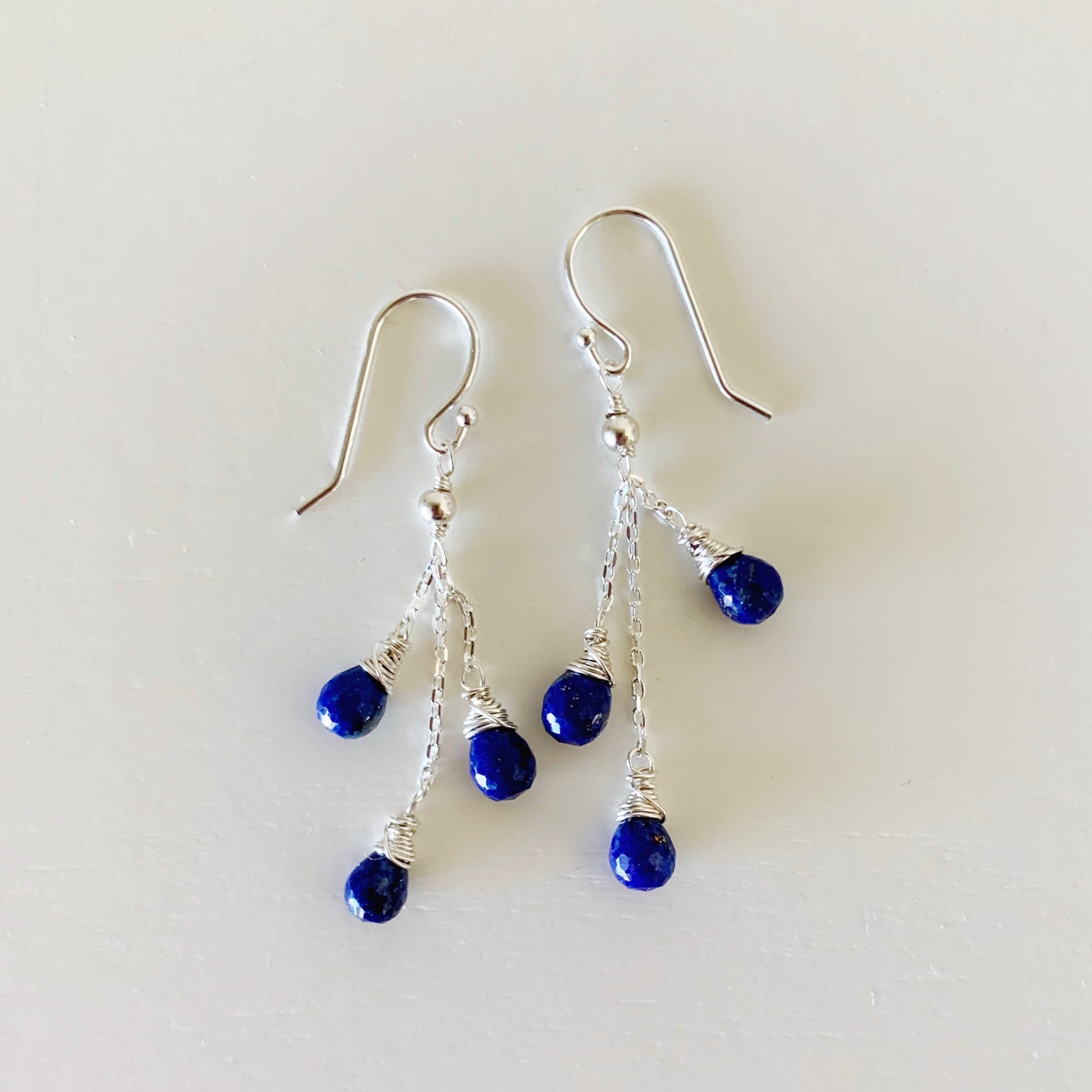 the neptune earrings by mermaids and madeleines are an eclectic style of earrings. these have three strands of sterling silver strands hanging from the earring hook with a royal blue lapis teardrop bead suspended from the bottom of each chain. this pair is photographed with the strands spread out and flat on a white surface