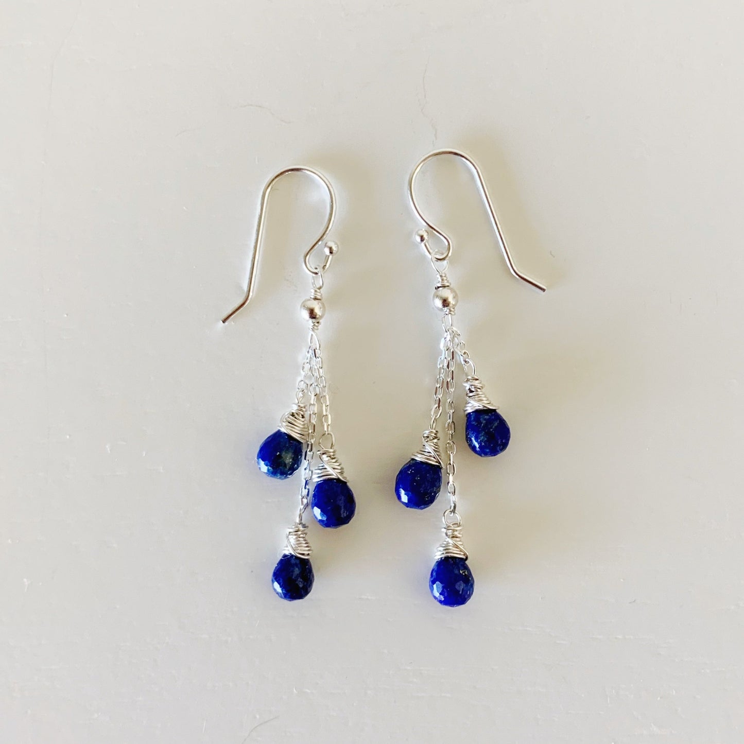 the neptune earrings by mermaids and madeleines are a eclectic style with three strands of sterling silver chain on each earring hook with royal blue lapis teardrop wire wrapped beads on the end of each strand. this pair of earrings is photographed flat on a white surface