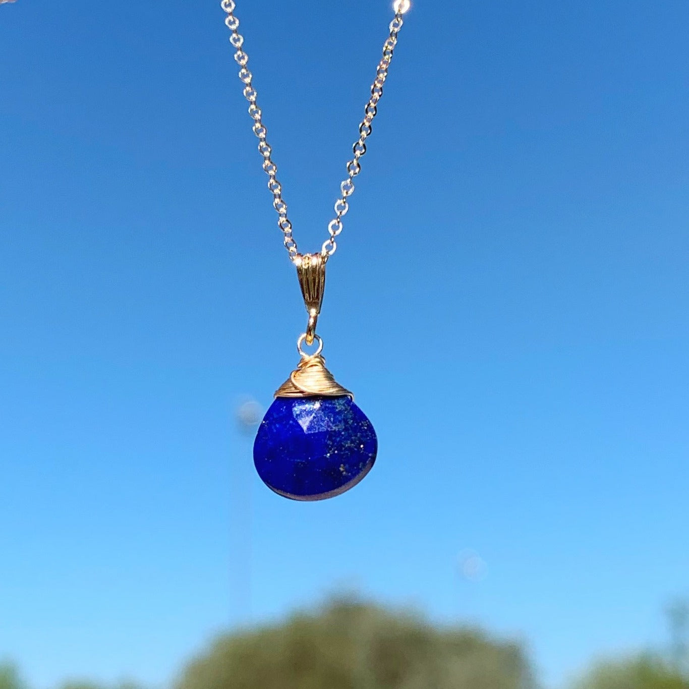 The neptune pendant necklace designed by mermaids and madeleines is created with blue lapis and 14k gold filled findings and chain. this necklace is suspended with blue sky in the background.