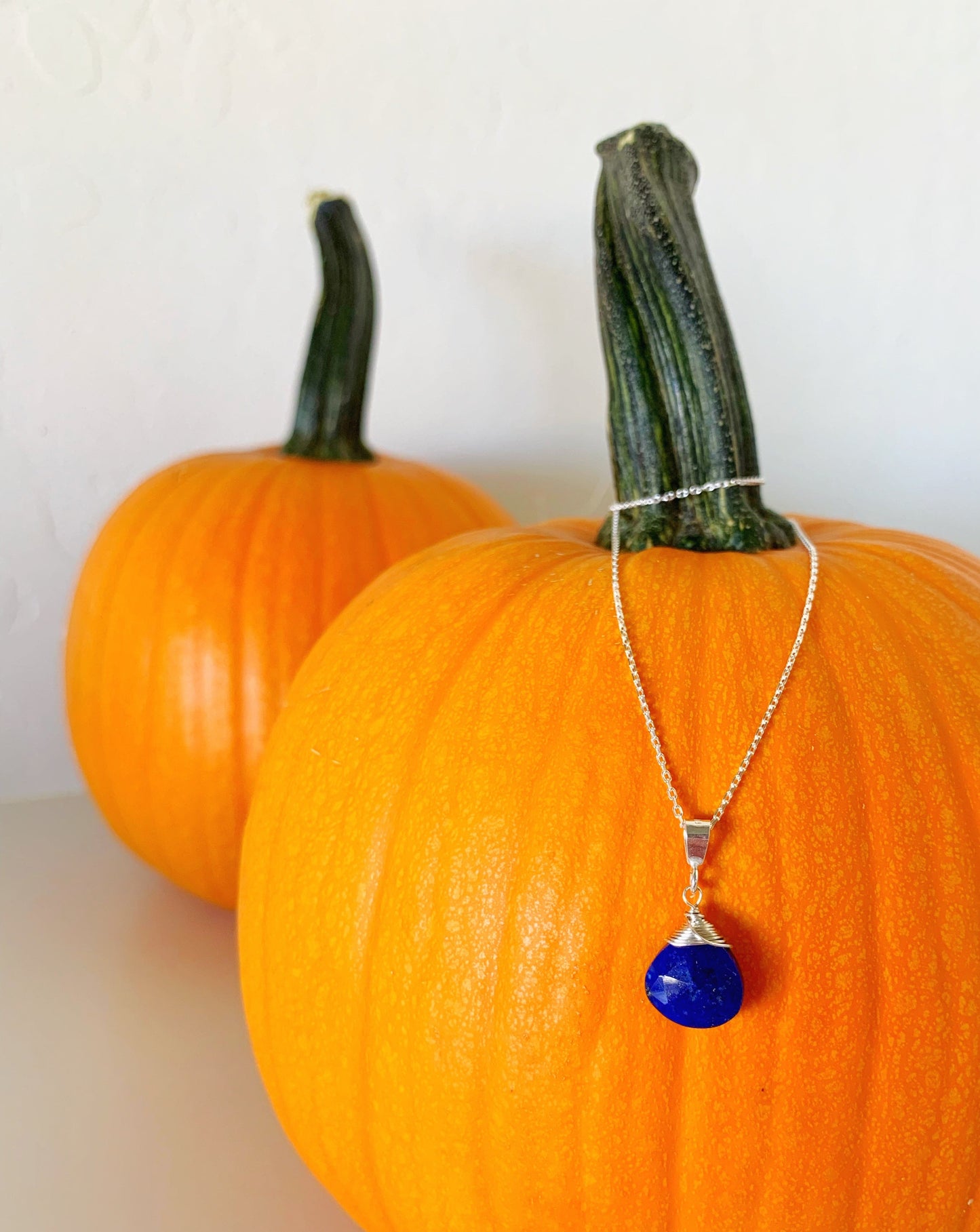 neptune necklace by mermaids and madeleines is a blue lapis faceted briolette wrapped with sterling silver on a sterling chain. this necklace is dangled off of a small pumpkin setting on a white surface