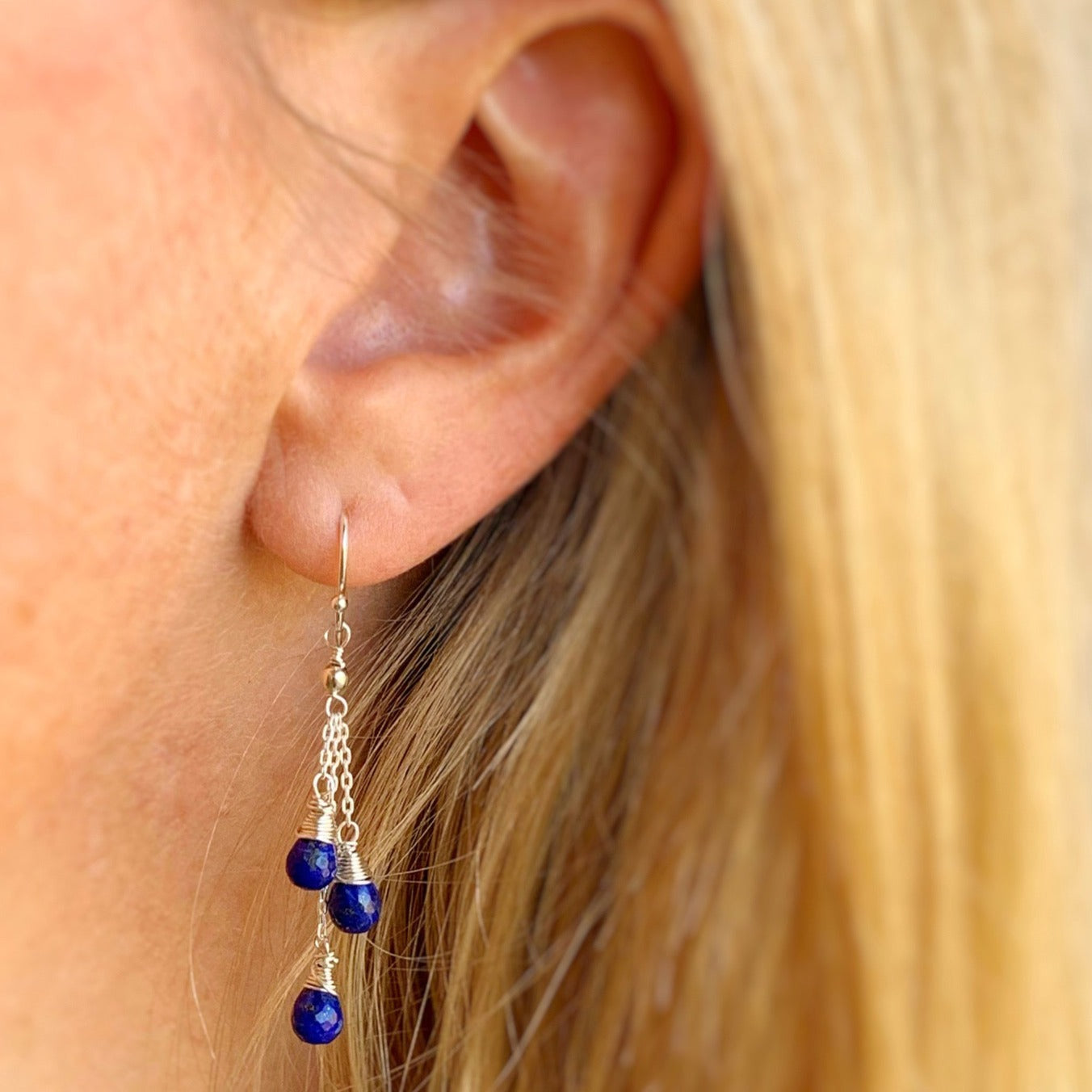 the neptune earrings by mermaids and madeleines are created with cascading lapis teardrop beads at 3 different lengths wire wrapped to sterling silver or 14k gold filled chain. the earring here is sterling silver and photographed worn on an ear and photographed close up.