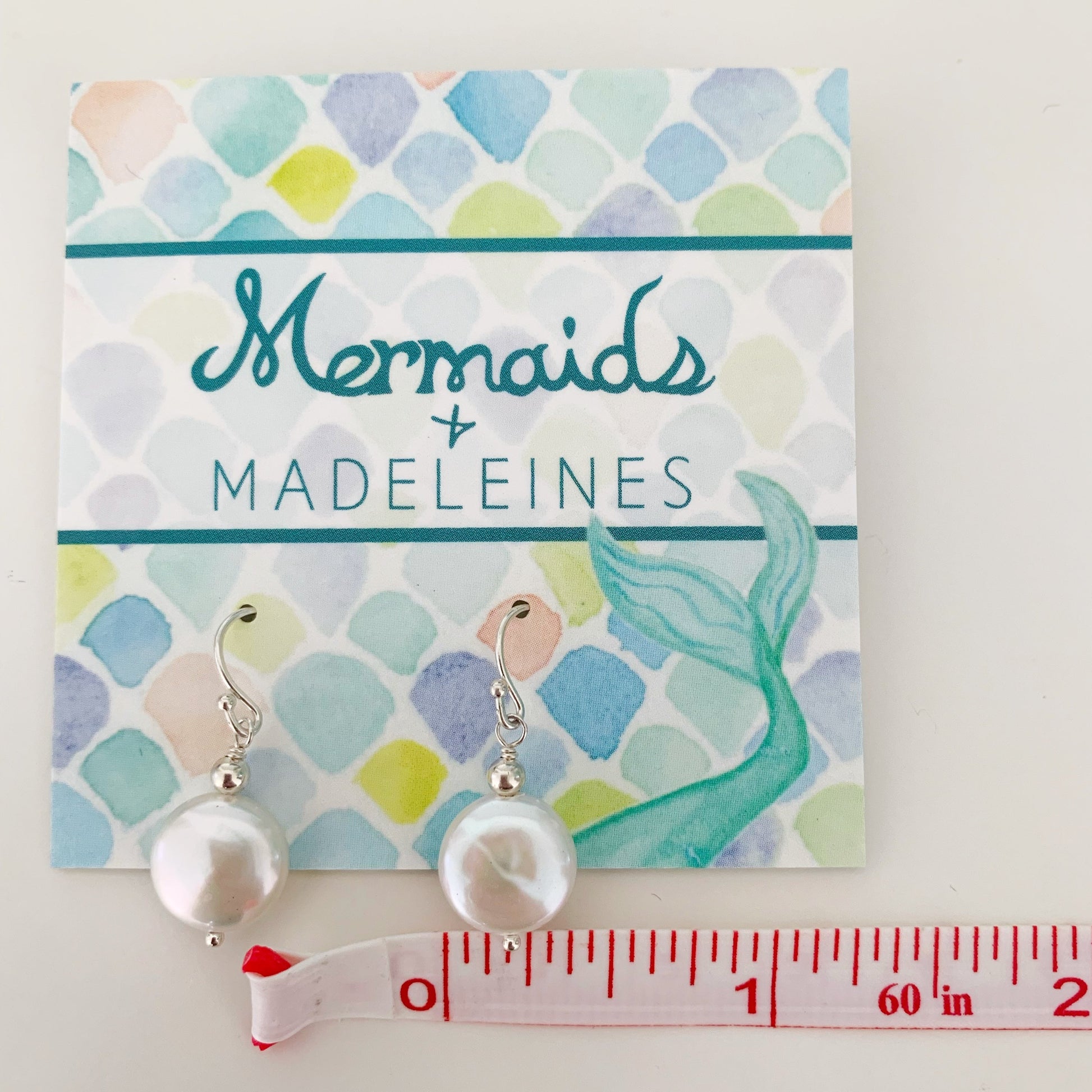 the newport earrings by mermaids and madeleines are freshwater coin pearl drops with sterling silver beads and findings. this pair is hanging on an earring card with a measuring tape near the pearl to show the pearl diameter of about 3/8". everything is photographed on a white surface
