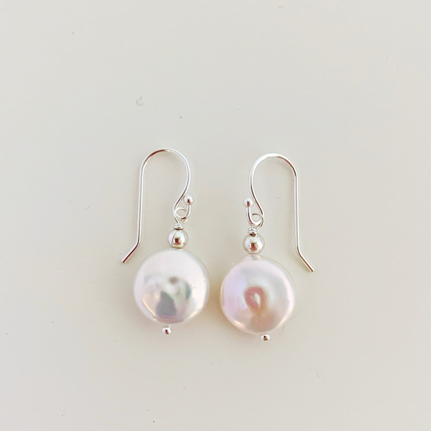 the newport earrings by mermaids and madeleines are a simple style earring designed with white, freshwater coin pearls and sterling silver findings and beads. this pair is photographed flat on a white surface