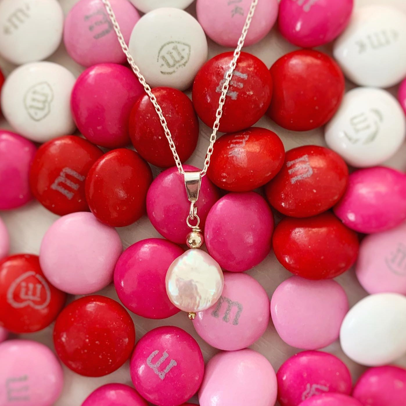 newport pendant necklace by mermaids and madeleines is created with a freshwater coin pearl hanging from sterling silver chain. this necklace is photographed on a bunch of red, pink, and white m&m candies