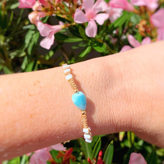 the ocean lover macrame bracelet by mermaids and madeleines is an adjustable friendship style bracelet with a larimar heart bead at the center and white opal chip beads on the sides and caramel color cord with a 14k gold filled slider clasp. this bracelet is photographed close up on a wrist in front of a pink flowered bush.