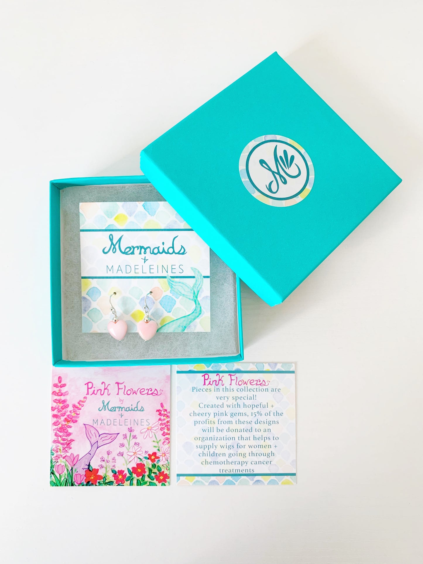 the pink posy earrings by mermaids and madeleines are light pink queen conch shells carved into a heart shape and complimented by a sterling silver bead on top and sterling silver findings. this pair is carded and placed in a teal gift box. in front of the box is a the story card that comes with the pieces in the pink flower collection. All items are photographed on a white surface.