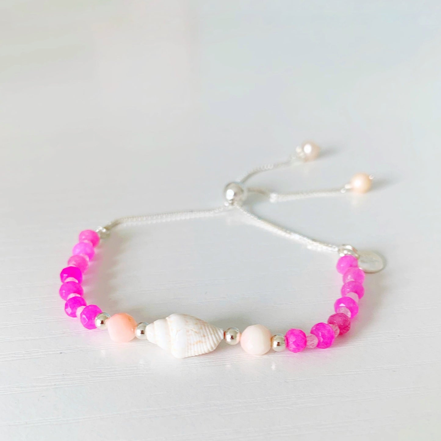 the pink flamingo shellebration bracelet by mermaids and madeleines is a sterling silver and beaded adjustable bracelet with a shell and coral beads at the center complimented by alternating dyed pink jade, rose quartz. this bracelet is photographed on a white surface