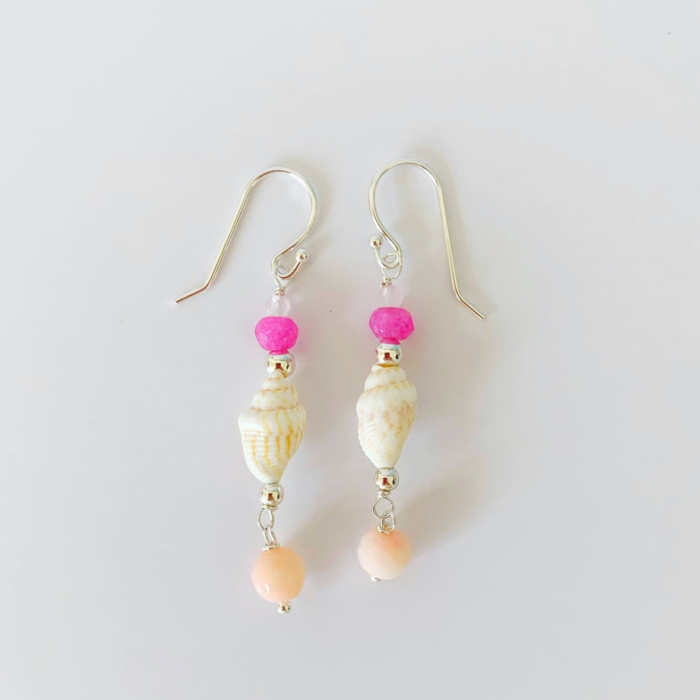 The pink flamingo shellebration earrings by mermaids and madeleines are a linear drop earring featuring a shell at the center complimented by dyed pink jade, rose quartz and a coral drop bead all with sterling silver findings. this pair is photographed on a white surface.