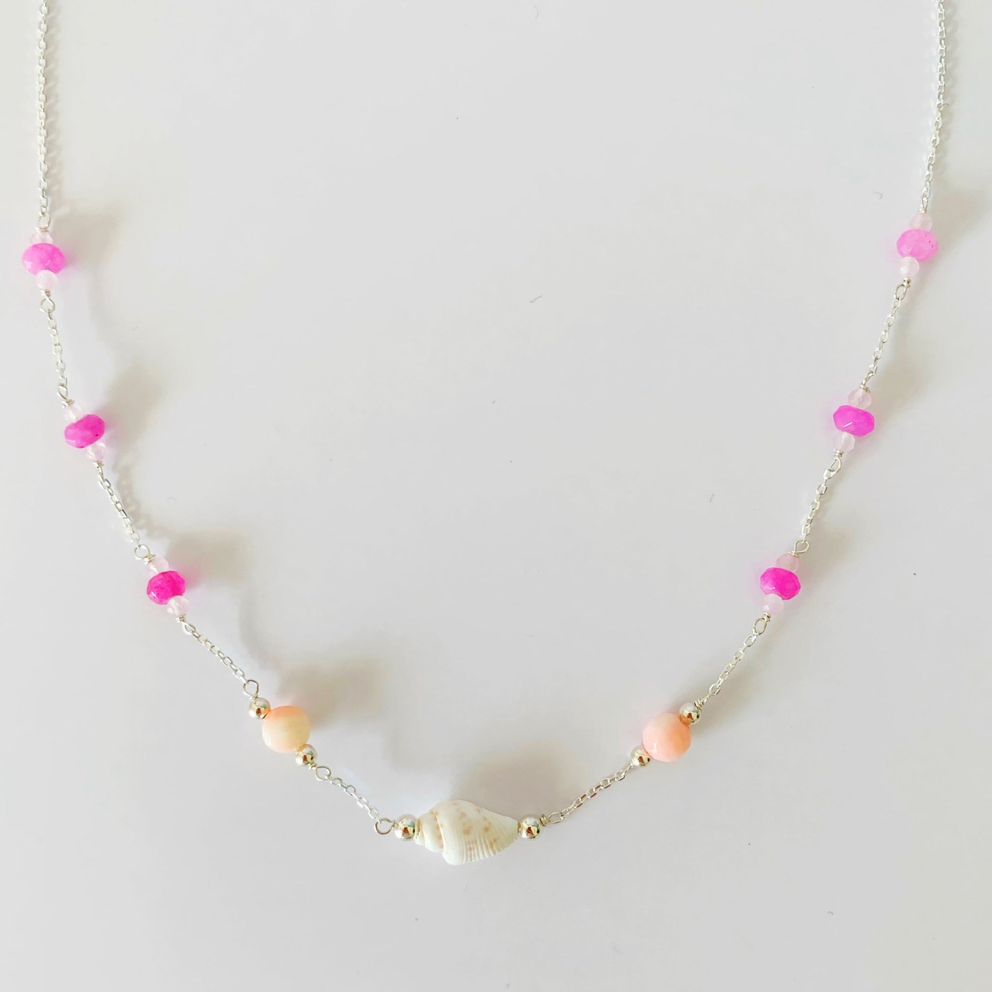 The pink flamingo shellebration necklace by mermaids and madeleines is created with a shell and coral beads at the center with dyed hot pink jade and rose quartz rondelles spaced out going up the sterling chain to the clasp. this necklace is photographed on a white surface