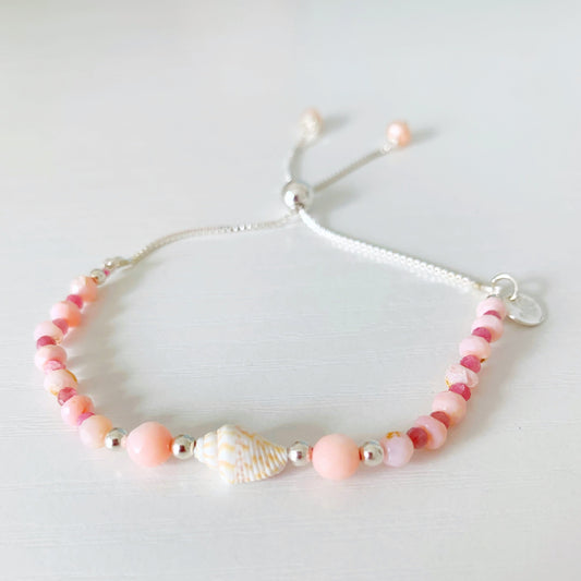 the pink lemonade shellebration bracelet by mermaids and madeleines is a sterling silver and beaded adjustable bracelet with a shell and coral beads at the center complimented by alternating pink opal and tourmaline. this bracelet is photographed on a white surface
