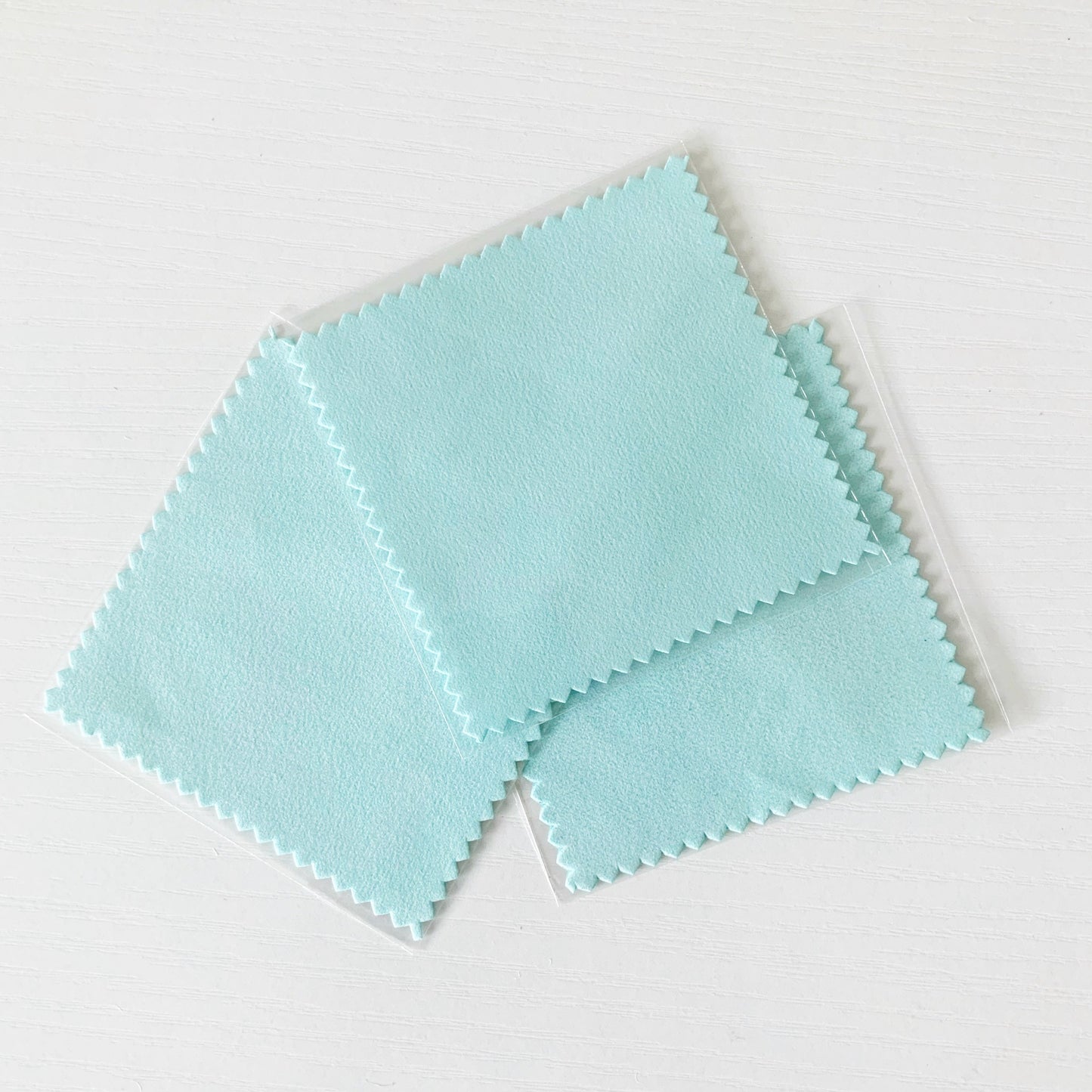 3 square light aqua color jewelry polishing cloths fanned out on a white surface