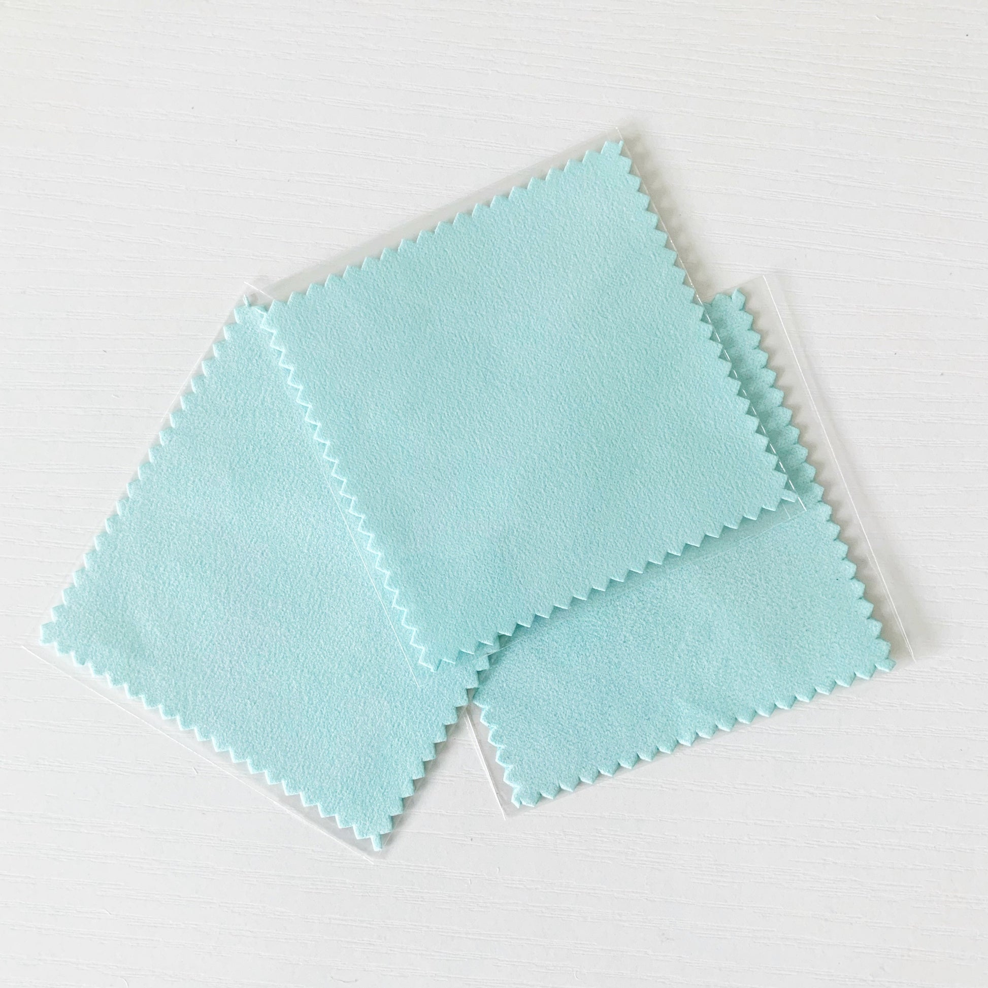3 square light aqua color jewelry polishing cloths fanned out on a white surface