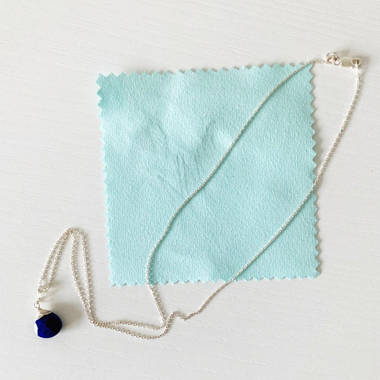 a light aqua jewelry polishing cloth with a few tarnish streaks on it and the mermaids and madeleine neptune necklace on top showing that the cloth can help renew the shine to sterling silver. all is photographed on white surface