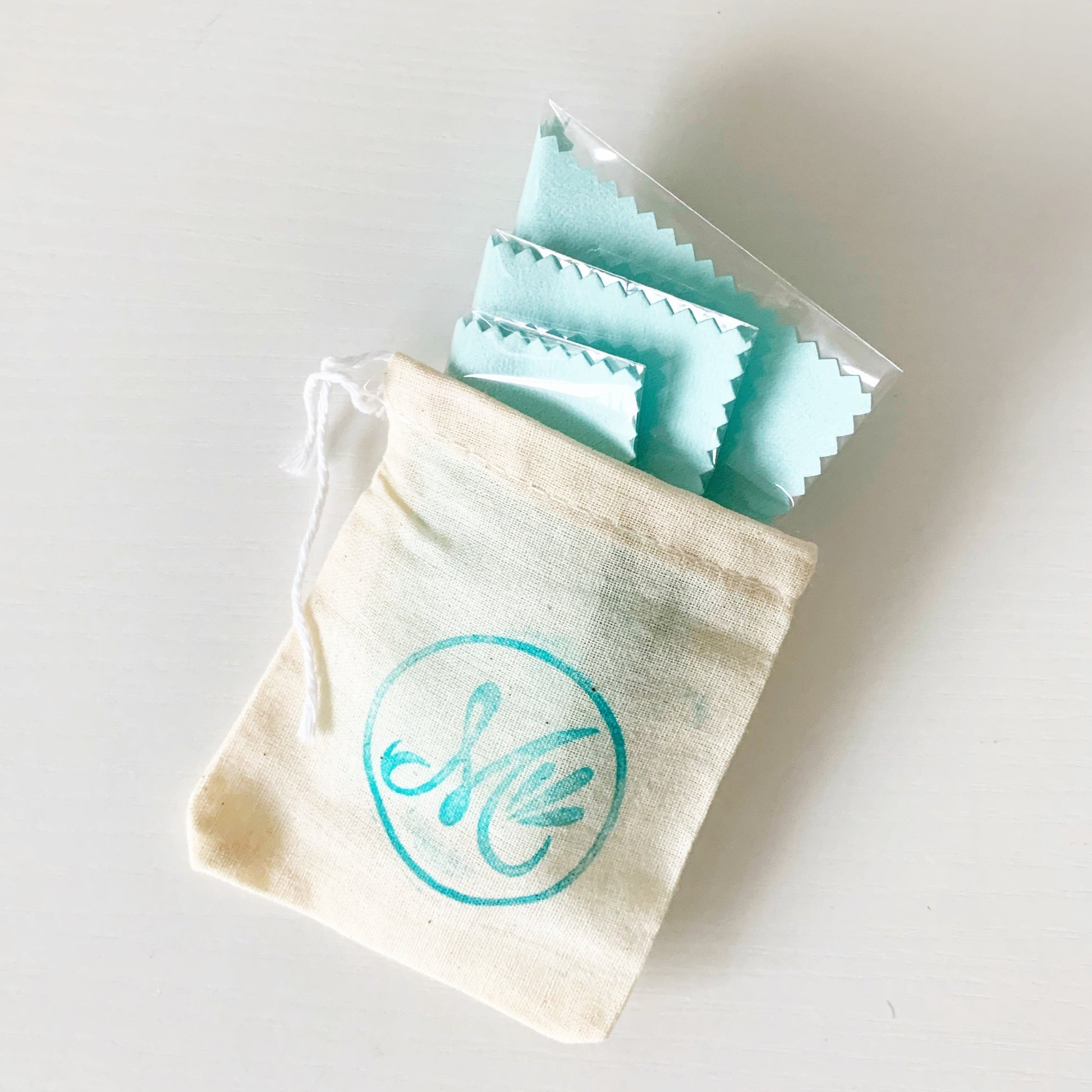 photograph of 3 light aqua jewelry polishing cloths folded into a mermaids and madeleines logo stamped cotton pouch. all photographed on a white surface