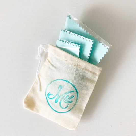 photograph of 3 light aqua jewelry polishing cloths folded into a mermaids and madeleines logo stamped cotton pouch. all photographed on a white surface