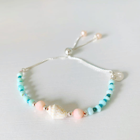 the pool party shellebration bracelet by mermaids and madeleines is a sterling silver and beaded adjustable bracelet with a shell and coral beads at the center complimented by alternating Peruvian opal and apatite. this bracelet is photographed on a white surface