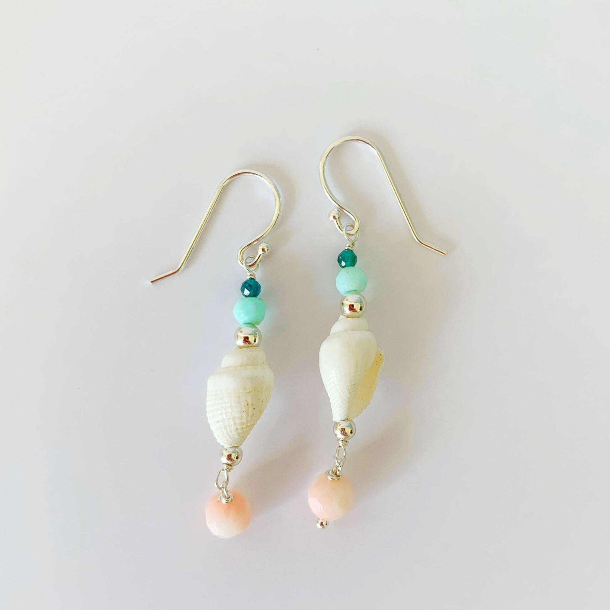 the pool party shellebration earrings by mermaids and madeleines are a linear drop style earrings with a shell at the center complimented by Peruvian opal, apatite, and with a coral bead drop at the bottom. this pair is photographed on a white surface