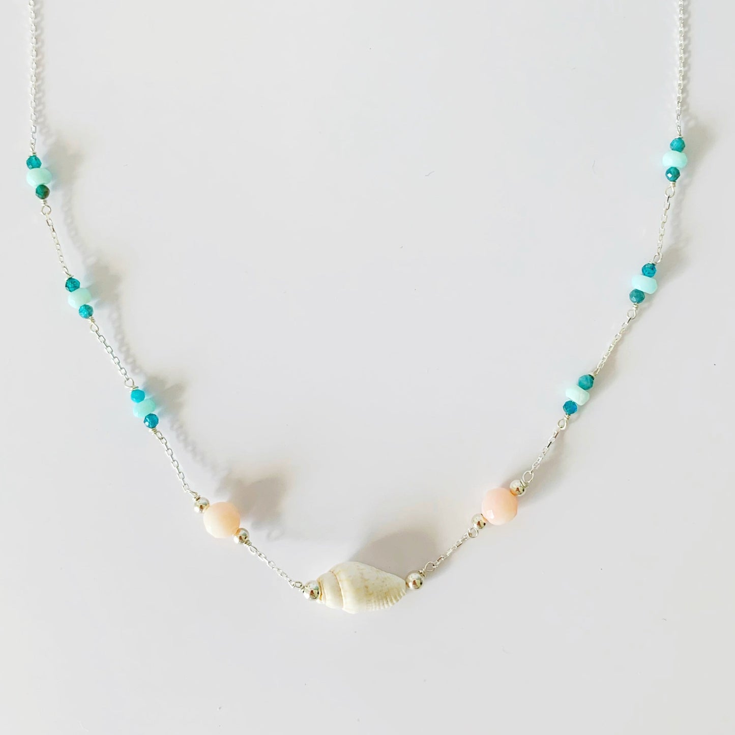 the pool party shellebration necklace by mermaids and madeleines is  station style necklace with a shell and coral beads at the center and little sections of Peruvian opal and apatite throughout the sterling chain to the clasp. this necklace is photographed on a white surface