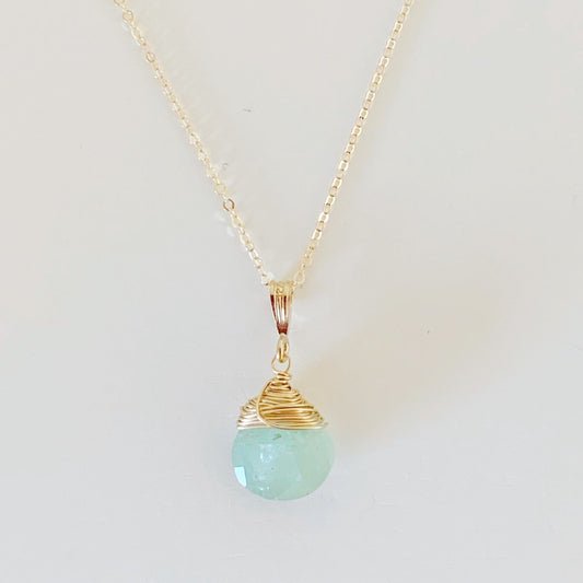 the raindrop necklace by mermaids and madeleines is a pendant style necklace featuring a ocean color aquamarine faceted briolette wire wrapped with 14k gold filled wire and hanging from a 14k gold filled chain. this necklace is photographed on a flat white surface