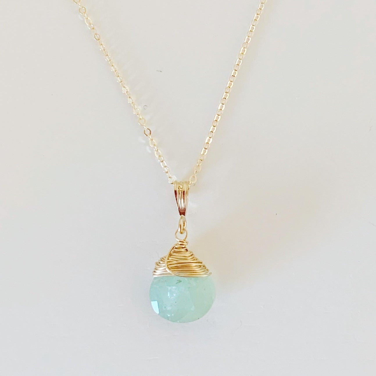 the raindrop necklace by mermaids and madeleines is a pendant style necklace featuring a ocean color aquamarine faceted briolette wire wrapped with 14k gold filled wire and hanging from a 14k gold filled chain. this necklace is photographed on a flat white surface