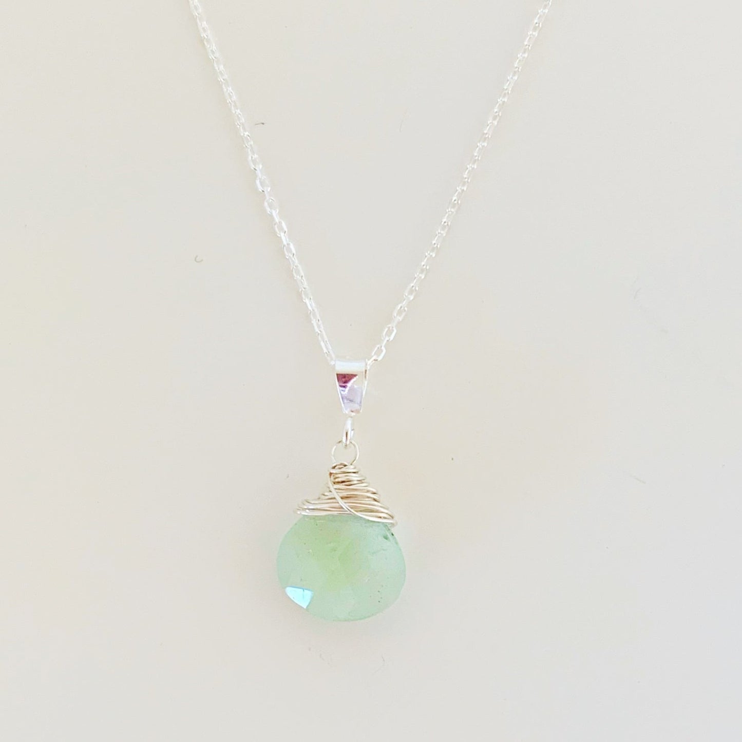 the raindrop necklace by mermaids and madeleines is a pendant design created with a faceted aquamarine briolette wire wrapped with sterling silver and hanging from sterling silver chain. photographed on a white surface