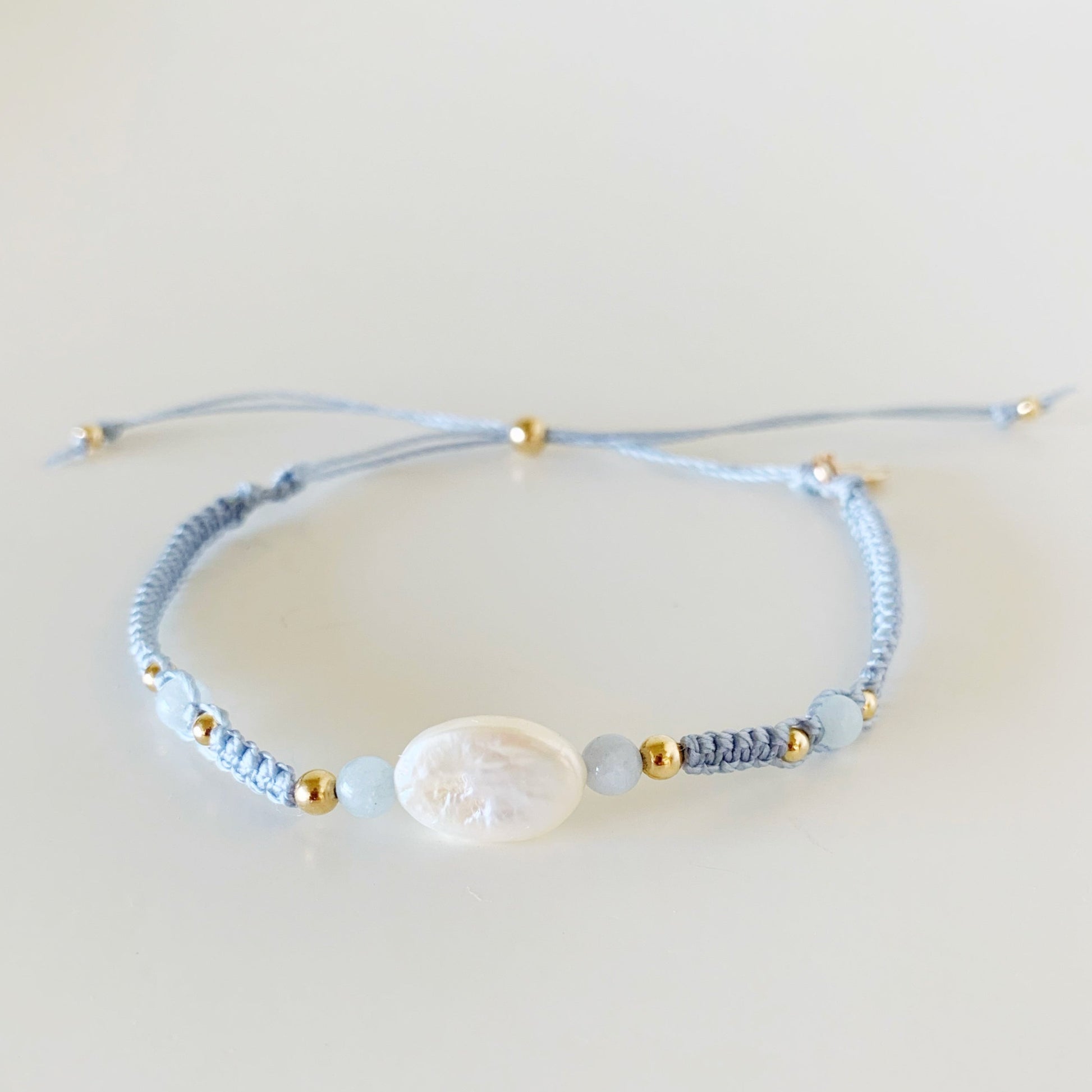 the regal seagull bracelet by mermaids and madeleines is an adjustable style macrame bracelet designed with blueish gray cord with a freshwater oval pearl at the center and 4mm aquamarine beads on either side. this bracelet is photographed from the front on a white surface