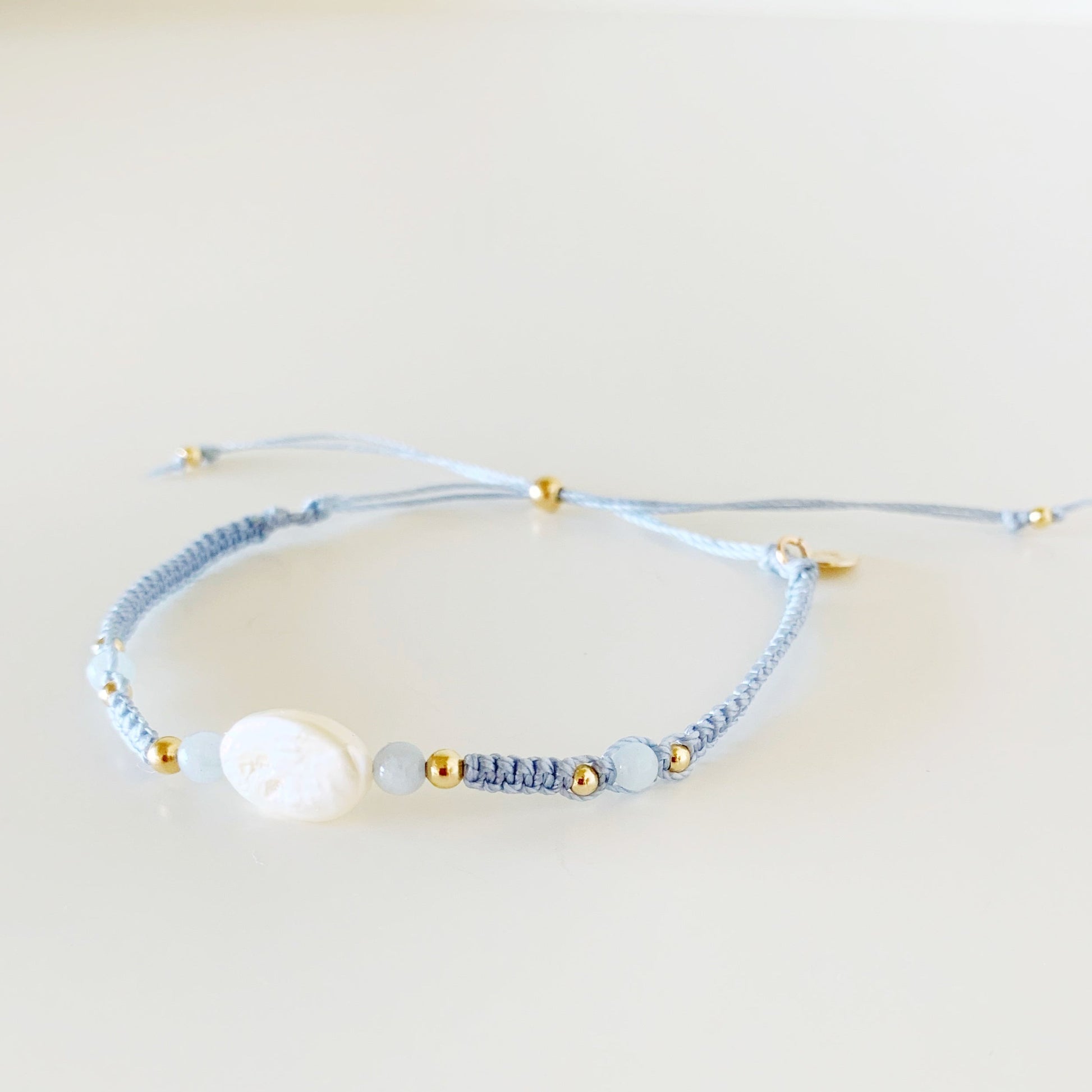 the regal seagull bracelet by mermaids and madeleines is an adjustable macrame bracelet with blue-gray cord with a freshwater oval pearl at the center with 4mm aquamarine beads on either side. this bracelet is photographed from a slight right angle with the piece on a white surface