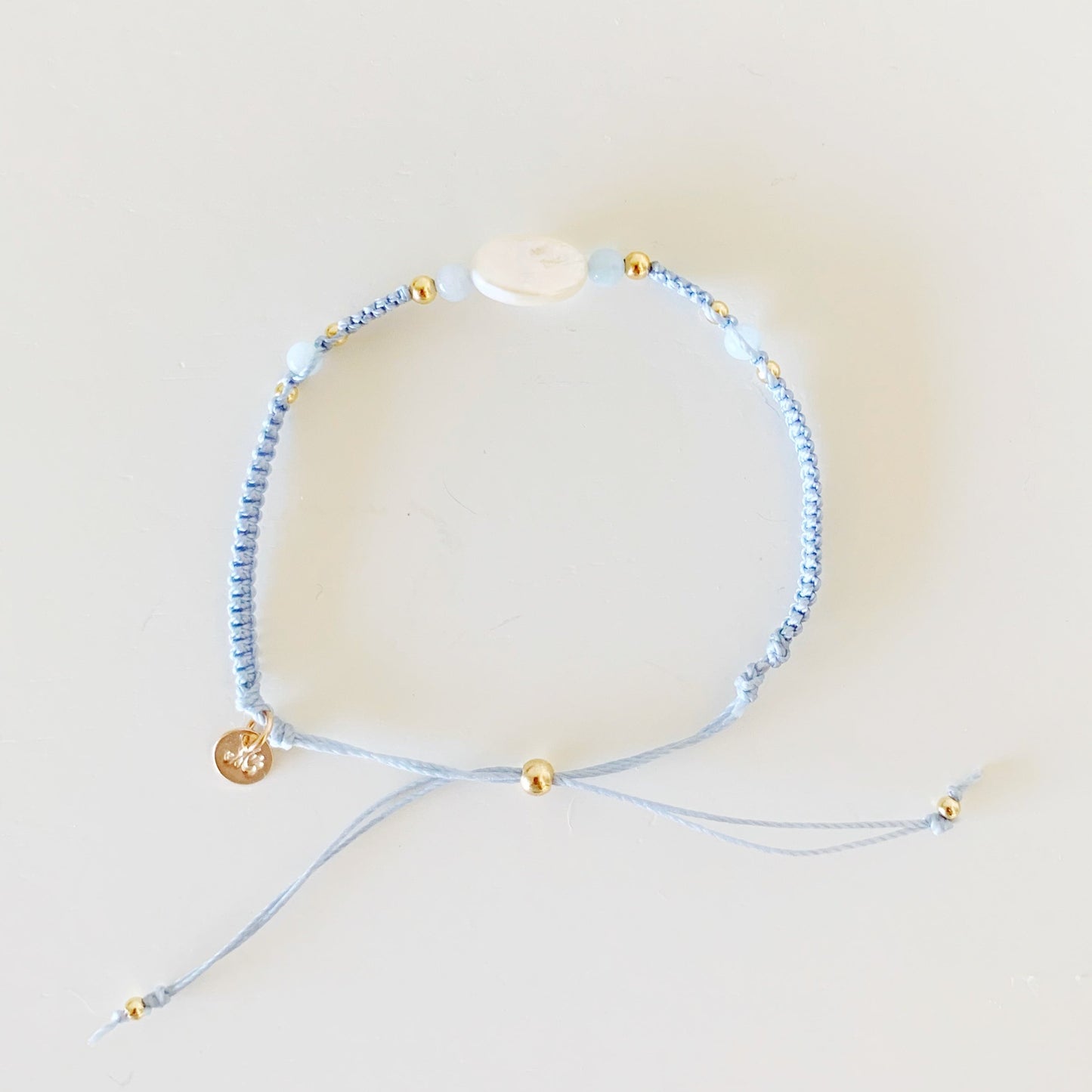 the regal seagull bracelet is a mermaids and madeleines design. its an adjustable macrame bracelet made with blue gray cord with a freshwater oval pearl at the center and aquamarine beads on both sides. this bracelet is photographed from the top down on a white surface