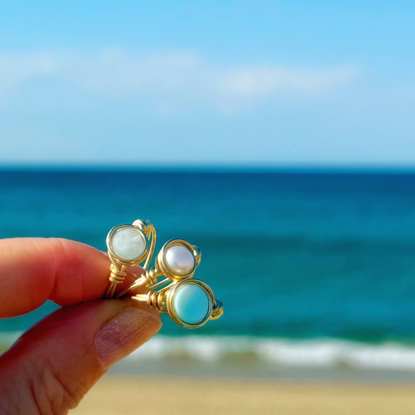 Gold filled wire-wrapped rings with gemstones held by a hand against a beach background