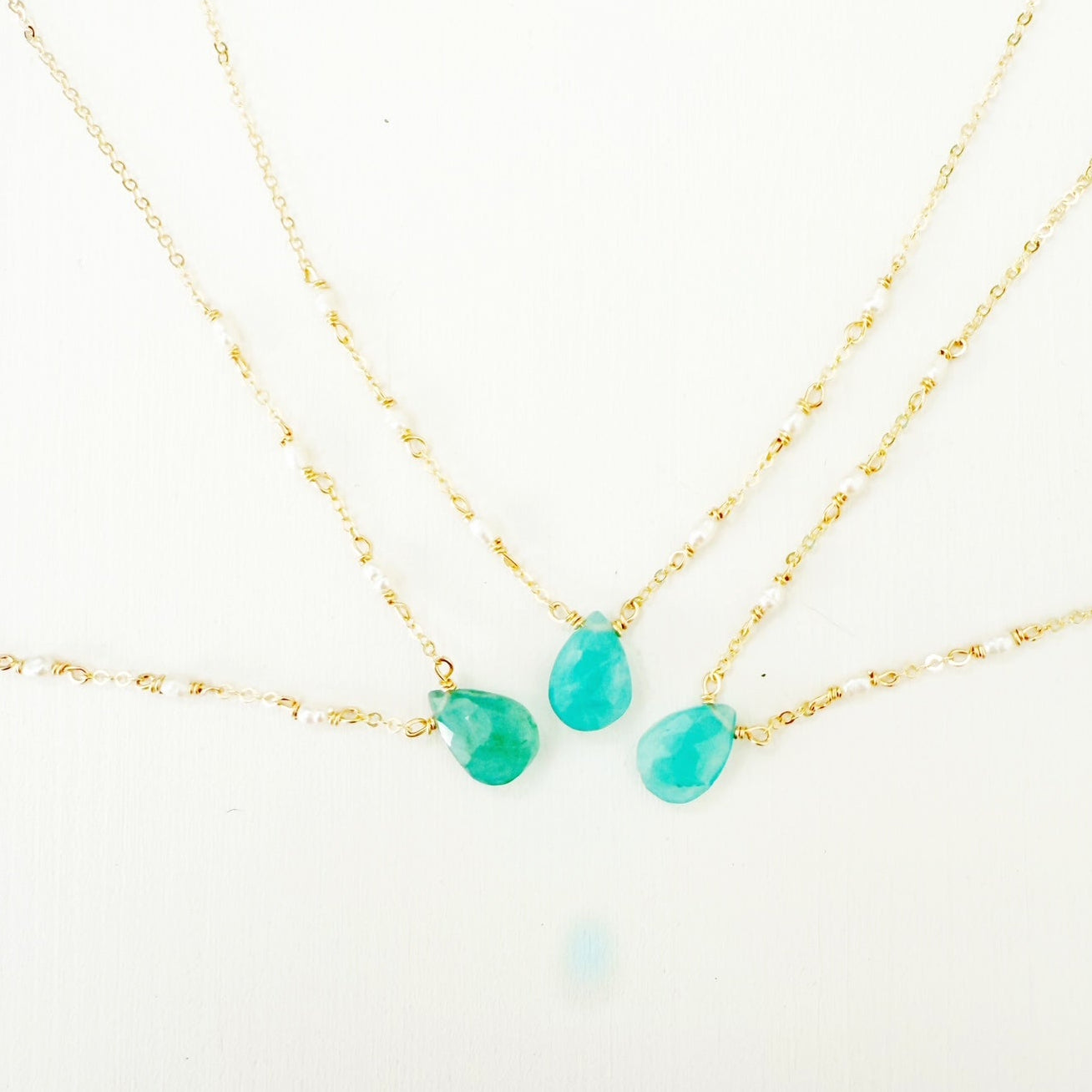 the sea salt necklace by mermaids and madeleines is a pendant style necklace on 14k gold filled chain with a grandidierite drop at the center and a few seed pearls on the chain on either side. this photo shows 3 of the necklaces close up on a white surface showing the range of color for the stone