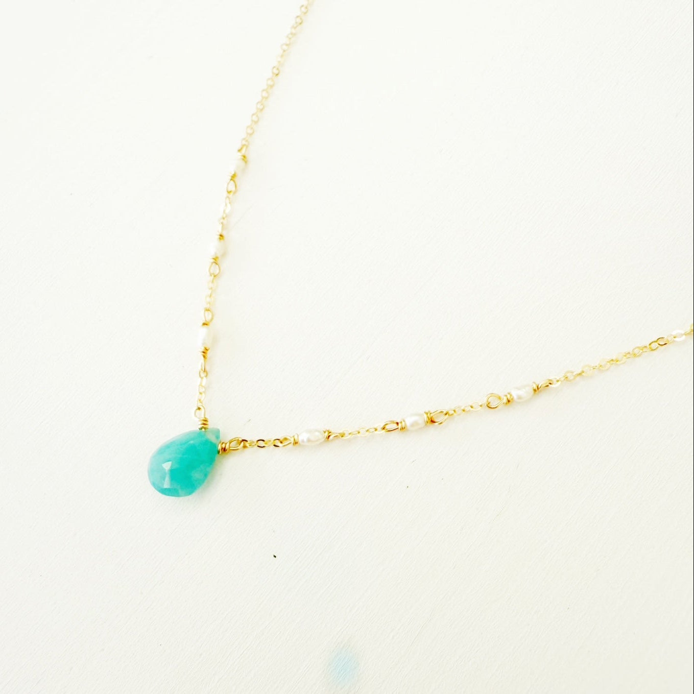 the sea salt necklace by mermaids and madeleines is a pendant style necklace on dainty 14k gold filled chain with a grandidierite drop at the center and seed pearls on either side. this is a close up side view of the necklace on a white surface