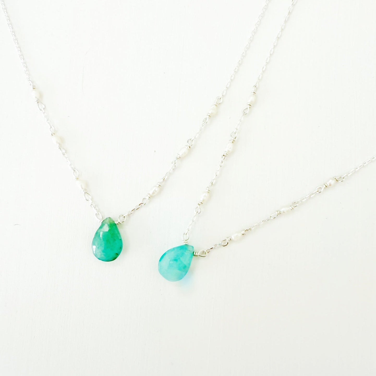 the sea salt necklace by mermaids and madeleines is a pendant style necklace with sterling silver chain and a grandidierite briolette at the center with a few seed pearls going up the chain on each side. this image shows  of these necklaces up close on a white surface